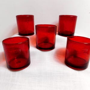 May include: Five red glass tumblers. The glasses are cylindrical with a slightly rounded base. The red color is consistent throughout the glass, and they are arranged on a white surface.