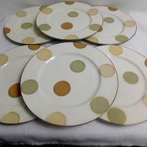 Puede incluir: Seis platos de cerámica blanca con borde marrón y un patrón de lunares grandes e irregulares en tonos marrón, verde y amarillo. Los platos están dispuestos sobre una superficie blanca.