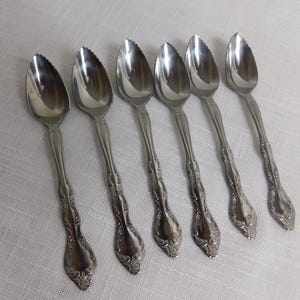 May include: Six silver grapefruit spoons arranged diagonally. Each spoon has a serrated edge on the bowl, a decorative handle, and a polished, reflective surface. The spoons are set against a white, textured background.