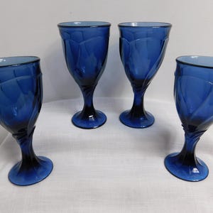 May include: Four cobalt blue stemmed water goblets. Each glass features a textured, petal-like design on the bowl and a dark blue stem and base. The glasses are arranged on a white surface.