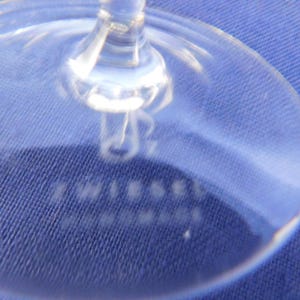 May include: Close-up of a clear wine glass base. The stem and base are visible, with the word "TWIESEL" and "HANDMADE" etched into the glass. The glass is on a blue textured surface.