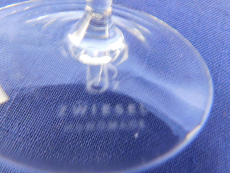 May include: Close-up of a clear wine glass base. The stem and base are visible, with the word "TWIESEL" and "HANDMADE" etched into the glass. The glass is on a blue textured surface.