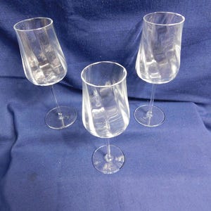 May include: Three clear wine glasses with a distinctive, twisted design. The glasses are made of transparent glass and are set against a blue background. The stems are slender, and the bases are round.