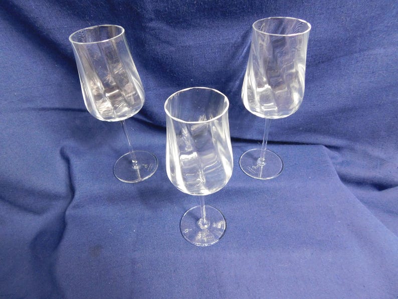 May include: Three clear wine glasses with a distinctive, twisted design. The glasses are made of transparent glass and are set against a blue background. The stems are slender, and the bases are round.