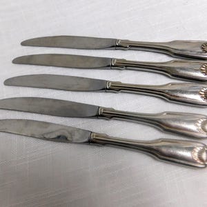 May include: Five silver butter knives with curved blades and decorative handles. The handles have a shell-like design at the end. The knives are arranged on a white surface.