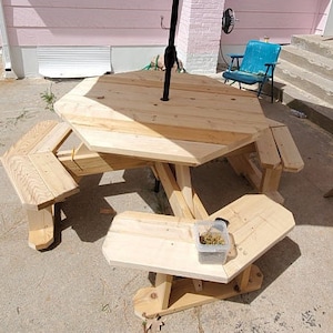 Op de afbeelding: Een achtekante houten picknicktafel met een beige parasol erboven. De tafel heeft drie banken die eraan vastzitten. De tafel is gemaakt van onbehandeld hout en staat op een betonnen terras.
