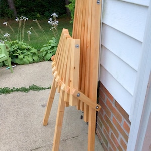 May include: A wooden folding chair with a natural finish. The chair is folded and leaning against a white wall with a brick base.
