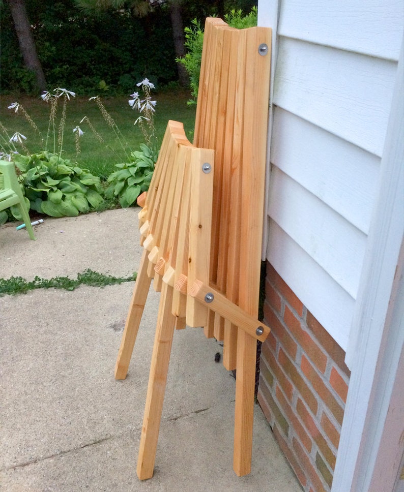 May include: A wooden folding chair with a natural finish. The chair is folded and leaning against a white wall with a brick base.