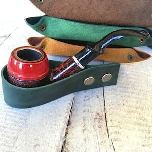 A green leather pipe holder with two brass buttons. A red and black pipe is resting in the holder.