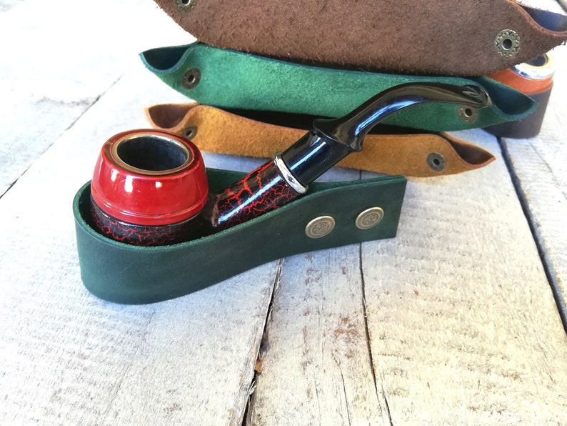 A green leather pipe holder with two brass buttons. A red and black pipe is resting in the holder.