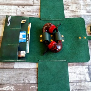 May include: A dark green leather roll-up tobacco pouch featuring a central holder for a tobacco pipe with a red and brown mottled bowl. The pouch includes side pockets containing a white lighter, a small metal tool, and a blue and white pack.