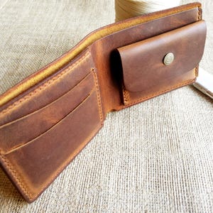 Könnte beinhalten: Ein braunes Leder-Bifold-Portemonnaie mit sichtbaren Nähten an den Kanten. Das Innere verfügt über mehrere Kartenfächer auf der linken Seite und ein kleines Münzfach auf der rechten Seite, das mit einem runden Metalldruckknopf verschlossen wird.