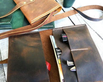 May include: Handmade leather journal in shades of brown, with a strap closure. The journal is open, revealing pockets and a pen. A green leather journal and a leather pen holder are also visible.