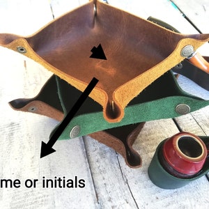 Three leather trays stacked on top of each other. The top tray is brown, the middle tray is green, and the bottom tray is brown. The trays are made of leather and have a stitched edge. The trays are designed to hold small items, such as jewelry, keys, or coins. The text 'Name or initials' is visible in the image.