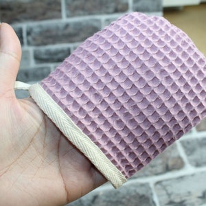 Könnte beinhalten: Ein weicher, rosa Waffelgewebe-Peelinghandschuh mit weißem Baumwollsaum. Der Handschuh ist so konzipiert, dass er an der Hand getragen und für eine sanfte Peeling verwendet werden kann.