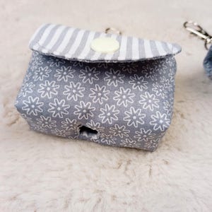 Könnte beinhalten: Kleine, graue Stofftasche mit weißem Blumenmuster und gestreifter Klappe. Die Tasche hat einen Knopfverschluss und einen silberfarbenen Schlüsselanhänger. Das Design ist schlicht und funktional, geeignet für kleine Gegenstände.