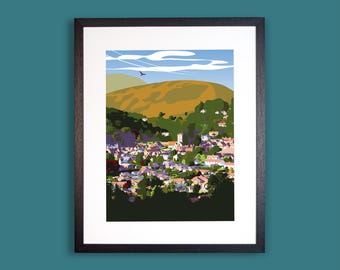 Könnte beinhalten: Ein gerahmter Kunstdruck mit einer stilisierten Landschaftsillustration einer Stadt am Fuße eines großen, goldbraunen Hügels unter einem hellblauen Himmel mit weißen Wolken. Ein kleiner dunkler Vogel ist am Himmel über dem Gelände zu sehen.