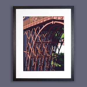 May include: Framed print of a section of a red iron bridge with intricate lattice and arch designs. The bridge is set against a dark blue background, with green foliage visible. The print is in a black frame.