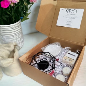 May include: A brown cardboard box filled with white shredded paper, a black metal candle holder, two white candles, a white ceramic dish, and a small white box with a floral design. The box is open and the contents are visible. A thank you note is visible on the inside of the box lid. The note says "Merci pour votre soutien. On veut vous voir avec vos bougies! Utilisez #scentricmood. Scantic."