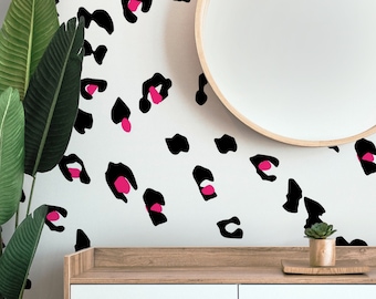 May include: White wall decorated with black and pink leopard print decals. A round wooden mirror hangs on the wall. A wooden cabinet with a plant and a small succulent in a pot are in the foreground.