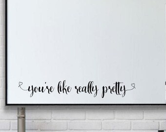 May include: A large rectangular mirror with a black frame is mounted above a gray vanity with three drawers. The mirror has the text "you're like really pretty" in a cursive font. The background is a white brick wall.