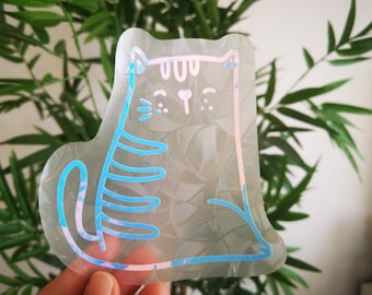 May include: A clear iridescent sticker with a blue outline of a cartoon cat. The cat has a pink nose and whiskers.
