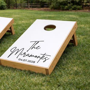 Könnte beinhalten: Ein Cornhole-Spielbrett aus Holz mit einer weißen Oberfläche und einem kreisförmigen Loch im oberen Bereich. Das Brett ist mit dem schwarzen Schriftzug The Miramontes und dem Datum 04.07.2028 in Schreibschrift bedruckt. Der Rahmen ist aus hellem Holz.