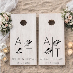Könnte beinhalten: Zwei helle Holz-Cornhole-Boards mit einem schwarzen Grafikdesign, bestehend aus den Buchstaben A und T, getrennt durch eine vertikale Linie und einen Blattzweig. Der Text lautet ABIGAIL & THOMAS und das Datum 17 | 5 | 20XX in schwarzer Schrift.