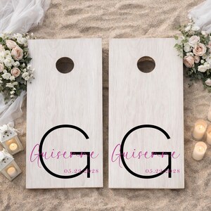 Könnte beinhalten: Ein Paar hellfarbige Holz-Cornhole-Boards mit einem großen schwarzen Buchstaben G, dem Namen Guiserne in pinker Schreibschrift und dem Datum 05.22.2028 in pinker Schrift. Jedes Board hat ein kreisförmiges Loch oben und eine helle Holzmaserung.