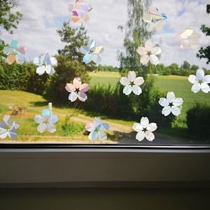 May include: Iridescent, flower-shaped window clings are adhered to a window screen. The clings have a multi-colored, rainbow-like sheen. The background shows a green field, trees, and a cloudy sky.