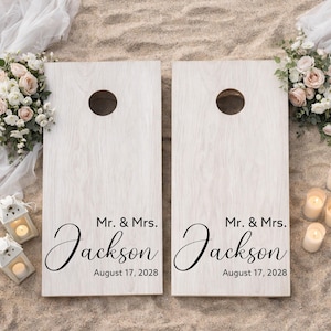 Könnte beinhalten: Zwei rechteckige Cornhole-Bretter mit heller Holzmaserung, die jeweils oben ein kreisförmiges Loch aufweisen. Die Bretter sind mit dem schwarzen Text Mr. & Mrs. Jackson und dem Datum August 17, 2028 in einer Kombination aus Serif- und Schreibschrift bedruckt.