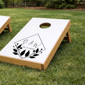 Könnte beinhalten: Ein weißes Cornhole-Spielbrett mit einem Rahmen aus Naturholz, das mit einer schwarzen Grafik in Rautenform versehen ist. Diese enthält die Buchstaben S & W, das Datum 22.5.2028 sowie ein Blatt- und Beerenmotiv. Oben befindet sich ein Loch.