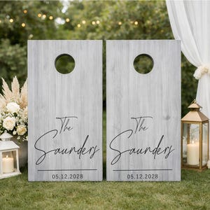 Könnte beinhalten: Zwei rechteckige Cornhole-Boards mit einer verwitterten grauen Holzmaserung. Jedes Board hat ein kreisförmiges Loch an der Oberseite und schwarzen Schreibschrifttext mit der Aufschrift The Saunders über dem Datum 05.12.2028 in serifenloser Schrift.