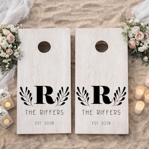 Könnte beinhalten: Zwei helle Holz-Cornhole-Boards mit einem schwarzen Buchstaben R, dem Text THE RIFFERS und dem Text EST. 2028. Jedes Board hat oben ein kreisförmiges Loch und ist mit schwarzen Blattillustrationen auf beiden Seiten des Buchstabens verziert.