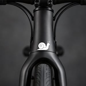 Peut inclure: Un cadre de vélo noir mat avec un autocollant d'escargot blanc sur le tube de direction. Le vélo comprend une fourche avant, une potence et des câbles de frein noirs partant du guidon. Le pneu avant est visible au bas du cadre.