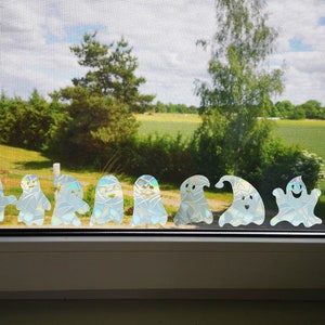 May include: A row of iridescent ghost stickers on a window, with a view of a green field and trees in the background.