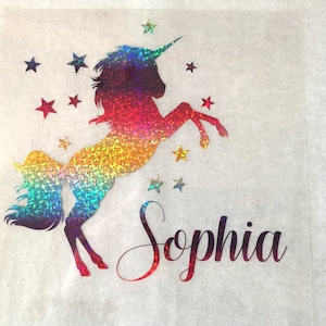Może przedstawiać: Jednorożec z tęczowego brokatu z rogiem i wzorem gwiazd. Jednorożec stoi na tylnych nogach z uniesionymi przednimi nogami. Nazwa "Sophia" jest napisana pod jednorożcem.