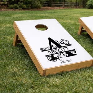 Könnte beinhalten: Ein weißes Cornhole-Board mit einem Rahmen aus Naturholz, das ein schwarzes Grafikdesign mit einem großen Buchstaben A zeigt, in dessen Mitte der Name ABBOTT steht, sowie den darunter gedruckten Text EST. 2027 und ein kreisförmiges Loch.