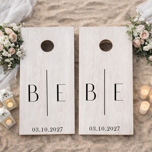 Könnte beinhalten: Zwei rechteckige Cornhole-Boards aus hellem Holz mit sichtbarer Maserung. Jedes Board hat ein kreisförmiges Loch oben, die Buchstaben B und E, die durch eine vertikale Linie getrennt sind, und das Datum 03.10.2027 unten aufgedruckt.