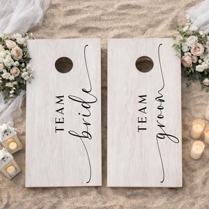 Könnte beinhalten: Zwei rechteckige Cornhole-Boards mit heller Holzmaserung. Jedes Board hat oben ein kreisförmiges Loch und schwarzen Text. Ein Board trägt die Aufschrift TEAM bride in Block- und Schreibschrift, das andere TEAM groom im gleichen Stil.
