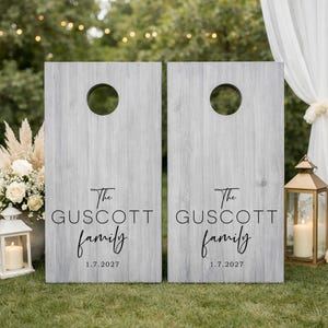 Könnte beinhalten: Zwei rechteckige Cornhole-Bretter mit einer hellgrauen Holzmaserung. Jedes Brett verfügt über ein kreisförmiges Loch an der Oberseite und schwarzen Text mit der Aufschrift The GUSCOTT family sowie das Datum 1.7.2027 in der Mitte.
