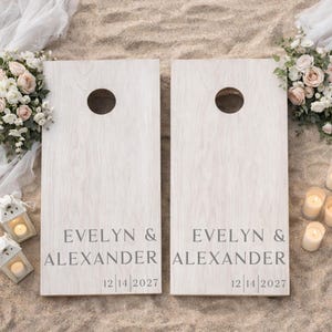 Könnte beinhalten: Zwei rechteckige Cornhole-Boards mit heller Holzmaserung, die jeweils oben ein kreisförmiges Loch aufweisen. Die Boards sind mit dem Text EVELYN & ALEXANDER und dem Datum 12 | 14 | 2027 in einer dunkelgrauen Serifenschrift bedruckt.