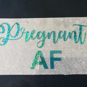 May include: A clear sticker with the words "Pregnant AF" in a teal, glittery font. The text is in a cursive style, with the letters "A" and "F" in a block style. The sticker is on a clear backing.