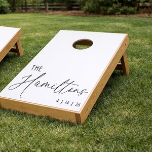 Könnte beinhalten: Ein weißes Cornhole-Spielbrett mit einem Rahmen aus Naturholz und klappbaren Beinen. Das Brett hat ein kreisförmiges Loch an der Oberseite und schwarzen Text mit der Aufschrift THE Hamiltons sowie dem Datum 4 | 14 | 26 in serifenloser Schrift.