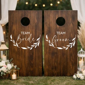 Könnte beinhalten: Zwei dunkelbraune hölzerne Cornhole-Boards mit einem kreisförmigen Loch im oberen Bereich. Jedes Board zeigt weißen Text und eine Blattgrafik. Das linke Board trägt die Aufschrift Team Bride, das rechte Team Groom in Schreibschrift.