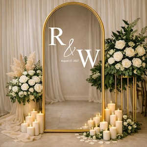 Peut inclure: Un grand miroir doré en forme d'arche, orné des initiales "R & W" et de la date "August 27, 2027" en blanc. L'ensemble est décoré de bougies blanches, de compositions florales et d'herbe de la pampa, créant une ambiance romantique et élégante.