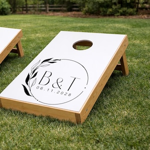 May include: A white cornhole board with a natural wood frame featuring a black circular graphic. The design includes a leafy branch, the letters B & T, and the date 06.11.2028 printed in black ink. The board has a circular hole at the top center.