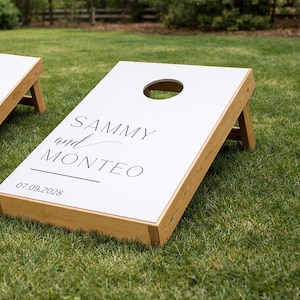 Könnte beinhalten: Ein Cornhole-Spielbrett aus Holz mit einer weißen Oberfläche, einem kreisförmigen Loch und schwarzem Aufdruck mit dem Text SAMMY and MONTEO sowie dem Datum 07.09.2028. Das Brett verfügt über einen Rahmen aus hellem Naturholz und Stützbeine.