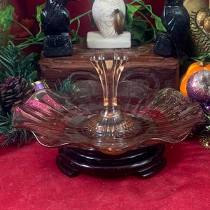 Puede incluir: Una exhibición de objetos decorativos sobre una superficie roja. Una fuente de vidrio rosa de dos niveles está en primer plano. Otros artículos incluyen una estatua blanca, figuras de cuervos negros y un tarro decorativo.