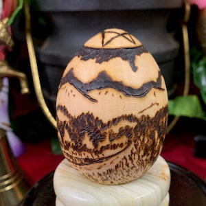 Może przedstawiać: Drewniana rzeźba w kształcie jaja z krajobrazem, w tym górami i drzewami, oraz pentagramem na górze. Jajko umieszczone jest na jasnej, warstwowej drewnianej podstawie i czarnej drewnianej podstawie.